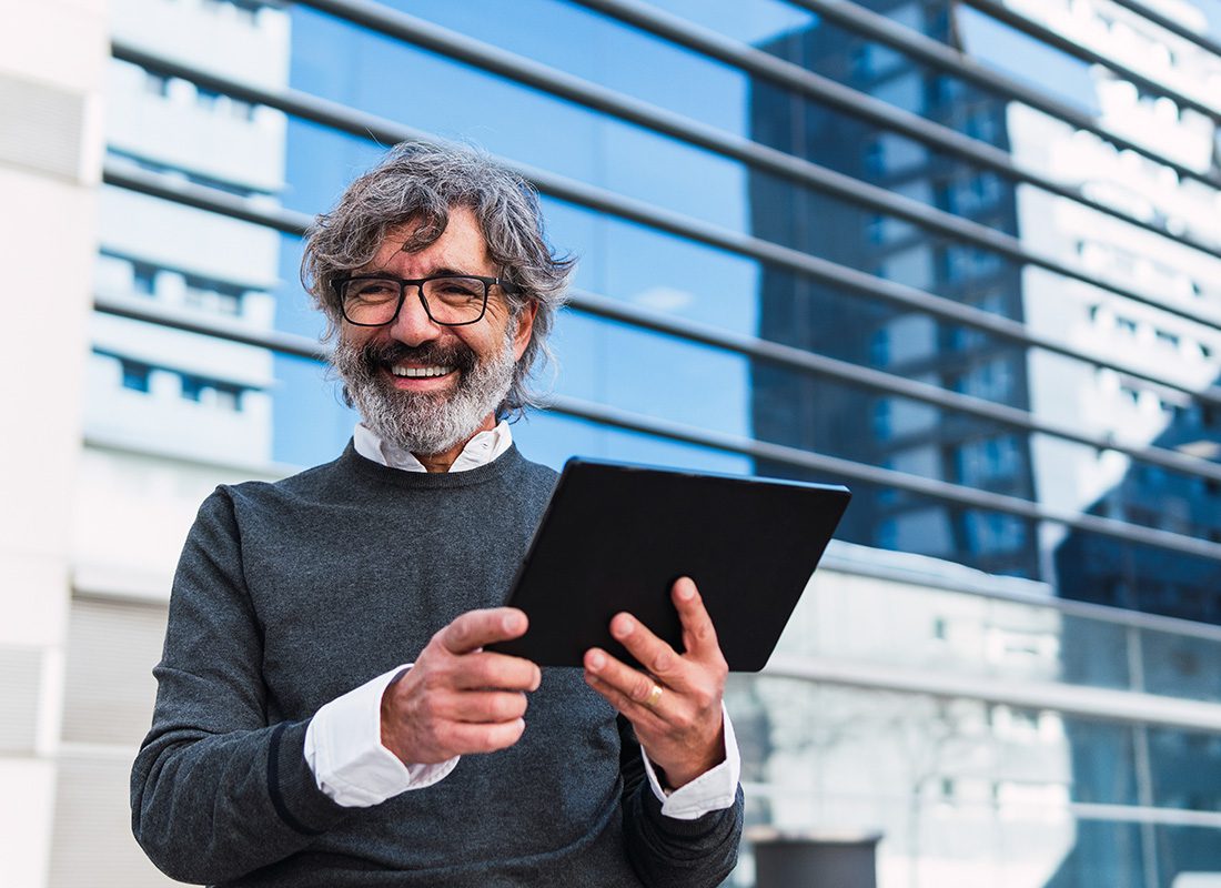 Business Insurance - Closeup Portrait of a Smiling Mature Businessman Standing Outside a Modern Commercial Office Building While Holding a Laptop in his Hands