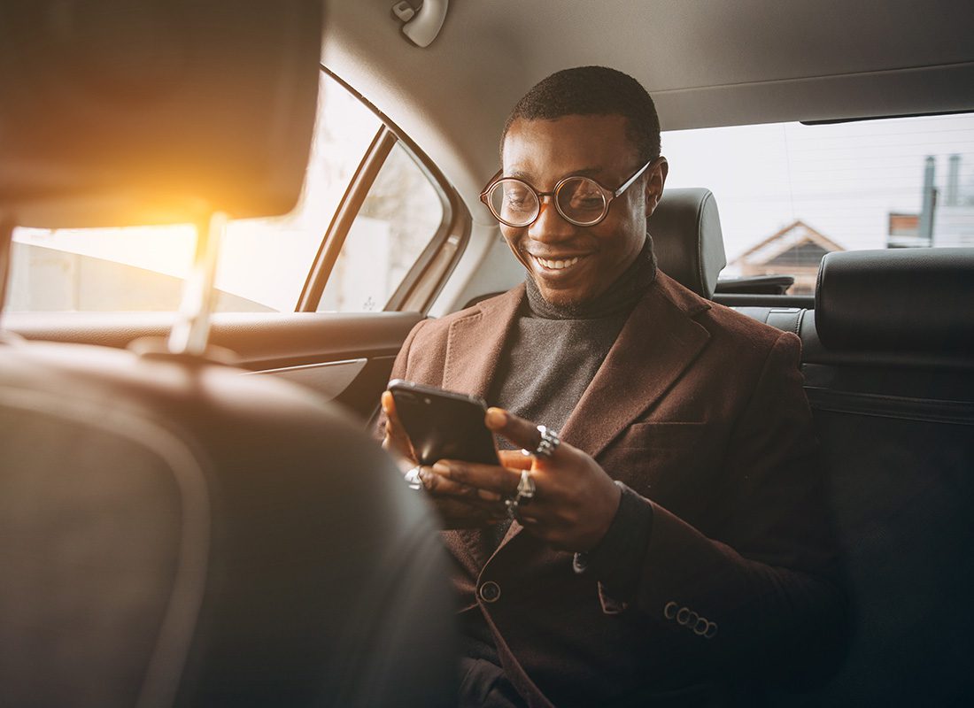 Service Center - Closeup View of a Smiling Young Man Wearing Glasses and a Blazer Looking at his Phone While Traveling in the Back Seat of a Car